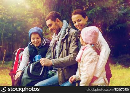 travel, tourism, hike and people concept - happy family sitting on bench and drinking hot tea from cups at camp in woods. happy family drinking hot tea at camp