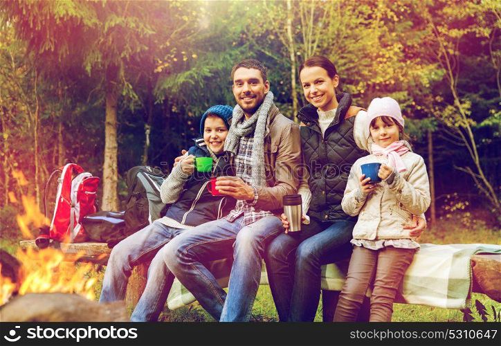 travel, tourism, hike and people concept - happy family sitting on bench and drinking hot tea from cups at camp fire in woods. happy family drinking hot tea at camp fire