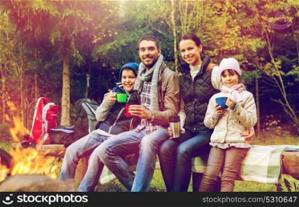 travel, tourism, hike and people concept - happy family sitting on bench and drinking hot tea from cups at camp fire in woods. happy family drinking hot tea at camp fire