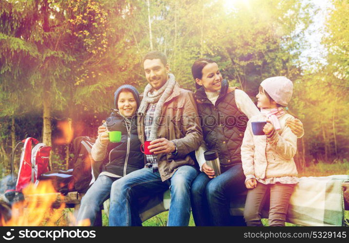 travel, tourism, hike and people concept - happy family sitting on bench and drinking hot tea from cups at camp fire in woods. happy family drinking hot tea at camp fire