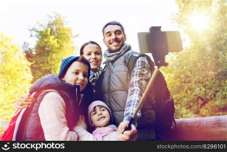 travel, tourism, hike and people concept - happy family with backpacks hiking and taking picture by smartphone on selfie stick. family with backpacks taking selfie and hiking