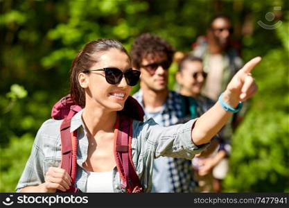 travel, tourism, hike and people concept - woman showing something to group of friends walking with backpacks in forest. group of friends with backpacks hiking in forest