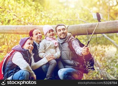 travel, tourism, hike and technoligy concept - happy family with backpacks taking picture by smartphone and selfie stick in woods. happy family with smartphone selfie stick in woods