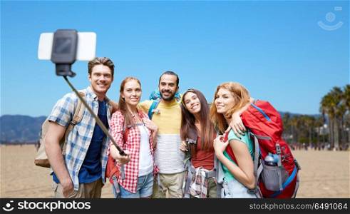 travel, tourism, hike and technology concept - group of smiling friends with backpacks taking picture by smartphone on selfie stick over venice beach background in california. friends with backpacks taking selfie by smartphone. friends with backpacks taking selfie by smartphone