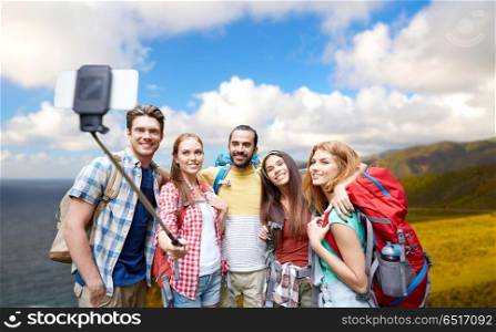 travel, tourism, hike and technology concept - group of smiling friends with backpacks taking picture by smartphone on selfie stick over big sur coast of california background. friends with backpacks taking selfie by smartphone. friends with backpacks taking selfie by smartphone