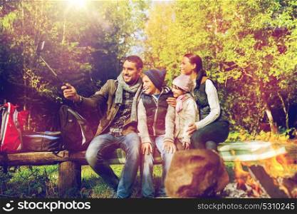 travel, tourism, hike and technology concept - happy family sitting on bench and taking picture with smartphone on selfie stick at campfire in woods. family with smartphone taking selfie near campfire