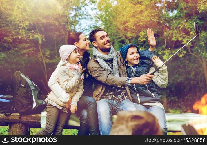 travel, tourism, hike and technology concept - happy family sitting on bench and taking picture with smartphone on selfie stick at campfire in woods. family with smartphone taking selfie near campfire