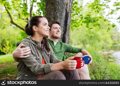 travel, tourism, hike, camping and people concept - happy couple with cups drinking tea in nature on river bank