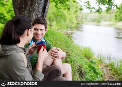 travel, tourism, hike, camping and people concept - happy couple with cups drinking tea in nature on river bank