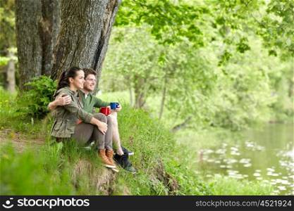 travel, tourism, hike, camping and people concept - happy couple with cups drinking tea and hugging in nature on river bank