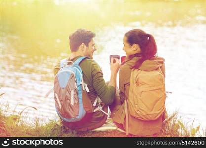 travel, tourism, hike, camping and people concept - happy couple with cups drinking tea in nature on river bank. happy couple with cups drinking in nature
