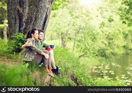 travel, tourism, hike, camping and people concept - happy couple with cups drinking tea and hugging in nature on river bank. happy couple with cups drinking tea in nature. happy couple with cups drinking tea in nature