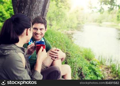 travel, tourism, hike, camping and people concept - happy couple with cups drinking tea in nature on river bank. happy couple with cups drinking in nature. happy couple with cups drinking in nature