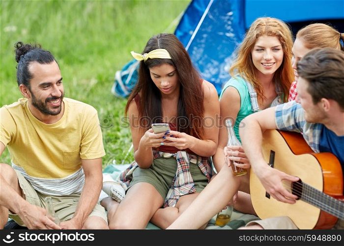 travel, tourism, hike, picnic and people concept - group of happy friends with smartphone and drinks playing guitar at camping