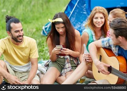 travel, tourism, hike, picnic and people concept - group of happy friends with smartphone and drinks playing guitar at camping