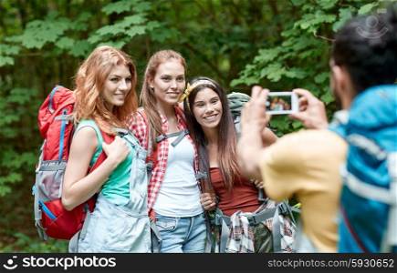 travel, tourism, hike, technology and people concept - group of smiling friends walking with backpacks taking picture by smartphone in woods