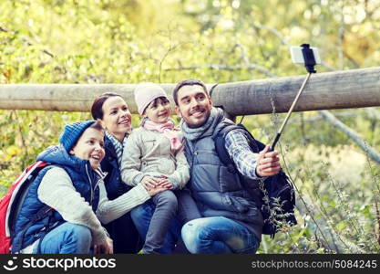 travel, tourism, hike, technology and people concept - happy family with backpacks taking picture by smartphone and selfie stick in woods. happy family with smartphone selfie stick in woods. happy family with smartphone selfie stick in woods