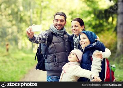 travel, tourism, hike, technology and people concept - happy family with backpacks taking selfie by smartphone in woods. family taking selfie with smartphone in woods. family taking selfie with smartphone in woods