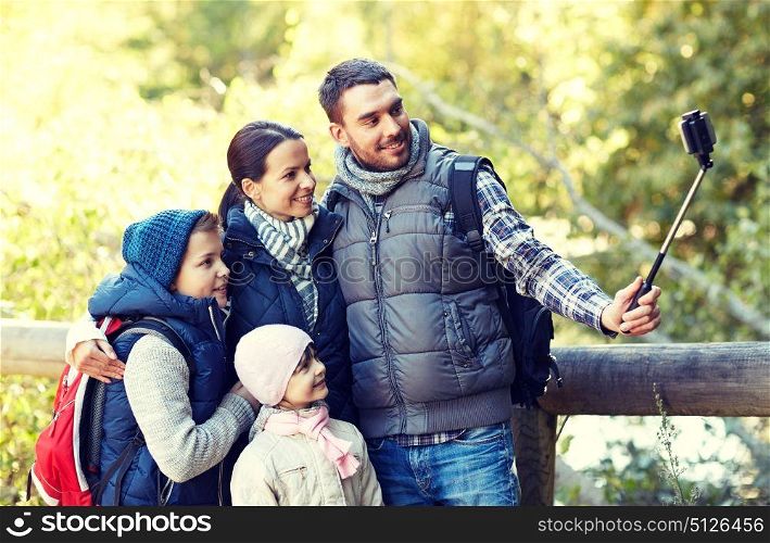 travel, tourism, hike, technology and people concept - happy family with backpacks taking picture by smartphone and selfie stick in woods. happy family with smartphone selfie stick in woods