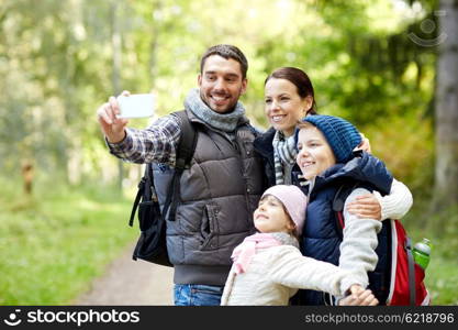 travel, tourism, hike, technology and people concept - happy family with backpacks taking selfie by smartphone in woods