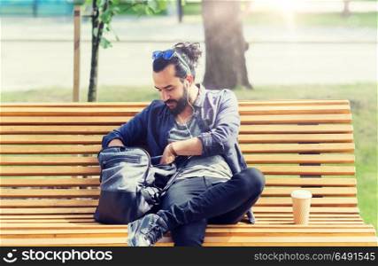 travel, tourism, lifestyle and people concept - man with earphones and coffee sitting on city bench and looking for something in his backpack. man with backpack and earphones in city. man with backpack and earphones in city