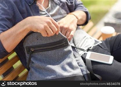 travel, tourism, lifestyle and people concept - man with earphones and sunglasses sitting on city bench and looking for something in his backpack