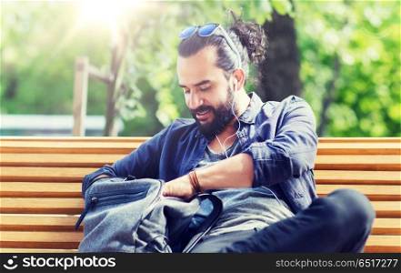 travel, tourism, lifestyle and people concept - man with earphones and sunglasses sitting on city bench and looking for something in his backpack. man with backpack and earphones in city. man with backpack and earphones in city