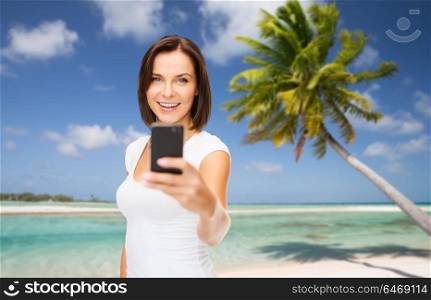 travel, tourism, summer holidays and vacation concept - happy young woman taking selfie by smartphone over exotic tropical beach with palm tree background. woman taking selfie by smartphone on beach