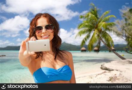 travel, tourism, summer holidays and vacation concept - happy young woman taking selfie by smartphone over exotic tropical beach with bungalow sheds background. woman taking selfie by smartphone on beach