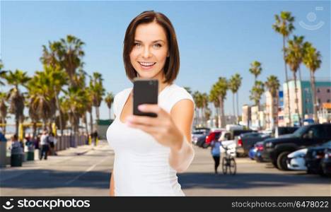 travel, tourism, summer holidays and vacation concept - happy young woman taking selfie by smartphone over venice beach in los angeles background. woman taking selfie by smartphone in los angeles. woman taking selfie by smartphone in los angeles