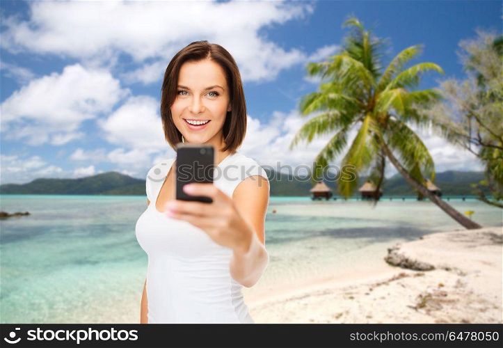 travel, tourism, summer holidays and vacation concept - happy young woman taking selfie by smartphone over exotic tropical beach with palm tree and bungalow sheds background in french polynesia. woman taking selfie by smartphone on beach. woman taking selfie by smartphone on beach