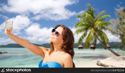 travel, tourism, summer holidays and vacation concept - happy young woman taking selfie by smartphone over exotic tropical beach with bungalow sheds background in french polynesia. woman taking selfie by smartphone on beach. woman taking selfie by smartphone on beach