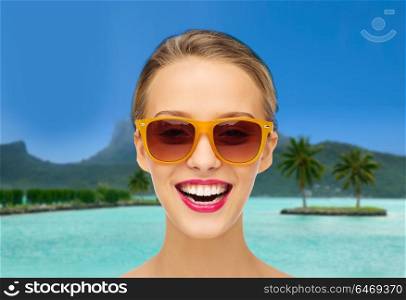 travel, tourism, summer holidays and vacation concept - smiling young woman in sunglasses with pink lipstick on lips over bora bora island beach background. woman in sunglasses over bora bora beach