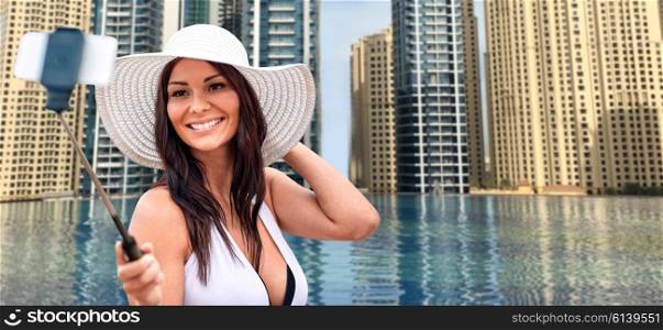 travel, tourism, summer, technology and people concept - smiling young woman in sun hat taking picture with smartphone on selfie stick over infinity edge swimming pool in dubai city background