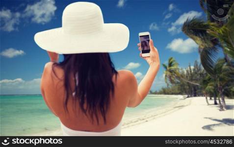 travel, tourism, summer, technology and people concept - smiling young woman or teenage girl in sun hat taking selfie with smartphone over tropical beach with palms background