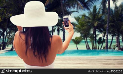travel, tourism, summer, technology and people concept - smiling young woman or teenage girl in sun hat taking selfie with smartphone over resort beach with palms and swimming pool background