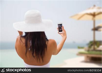 travel, tourism, summer, technology and people concept - smiling young woman or teenage girl in sun hat taking selfie with smartphone over resort beach with parasols and swimming pool background