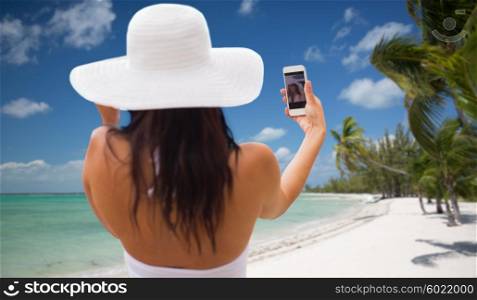 travel, tourism, summer, technology and people concept - smiling young woman or teenage girl in sun hat taking selfie with smartphone over tropical beach with palms background