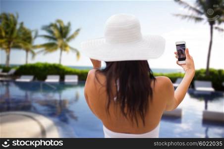 travel, tourism, summer, technology and people concept - smiling young woman or teenage girl in sun hat taking selfie with smartphone over resort beach with palms and swimming pool background