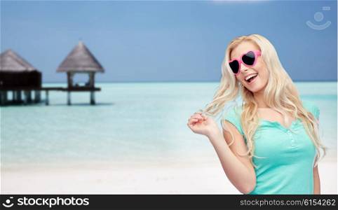 travel, tourism, summer vacation and people concept - smiling young woman or teenage girl in sunglasses holding her strand of hair over tropical beach with bungalow background
