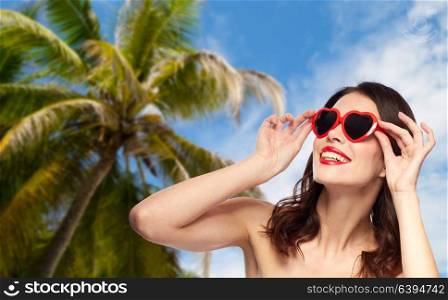 travel, tourism, summer vacation and valentines day concept - happy smiling young woman with red lipstick and heart shaped sunglasses over palm tree and blue sky background. woman with red lipstick and heart shaped shades