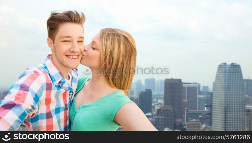 travel, tourism, summer vacation, technology and love concept - happy couple taking selfie with smartphone or camera and kissing over city background