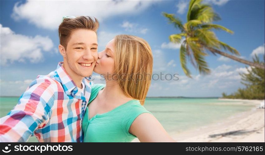 travel, tourism, summer vacation, technology and love concept - happy couple taking selfie with smartphone or camera and kissing over tropical beach background