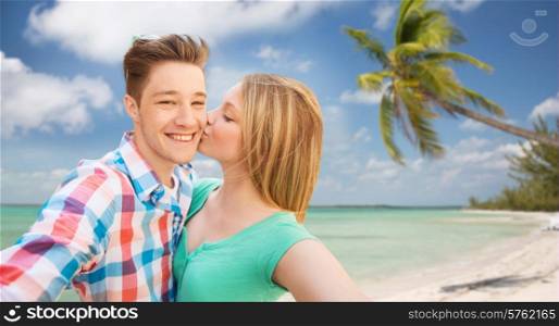 travel, tourism, summer vacation, technology and love concept - happy couple taking selfie with smartphone or camera and kissing over tropical beach background