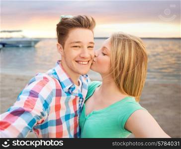 travel, tourism, summer vacation, technology and love concept - happy couple taking selfie with smartphone or camera and kissing over sunset on sea shore background
