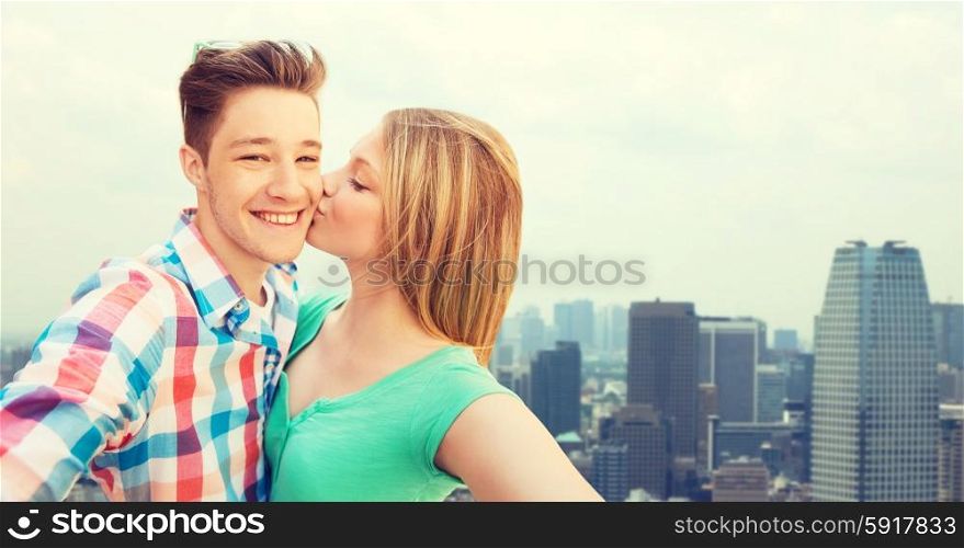 travel, tourism, summer vacation, technology and love concept - happy couple taking selfie with smartphone or camera and kissing over city background