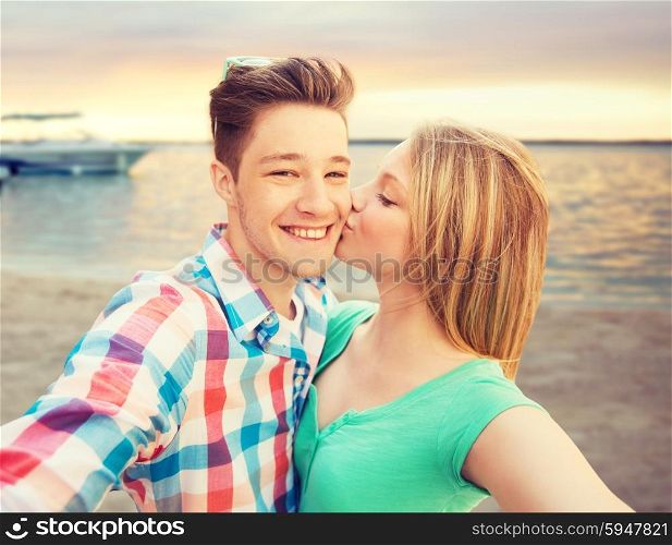 travel, tourism, summer vacation, technology and love concept - happy couple taking selfie with smartphone or camera and kissing over sunset on sea shore background