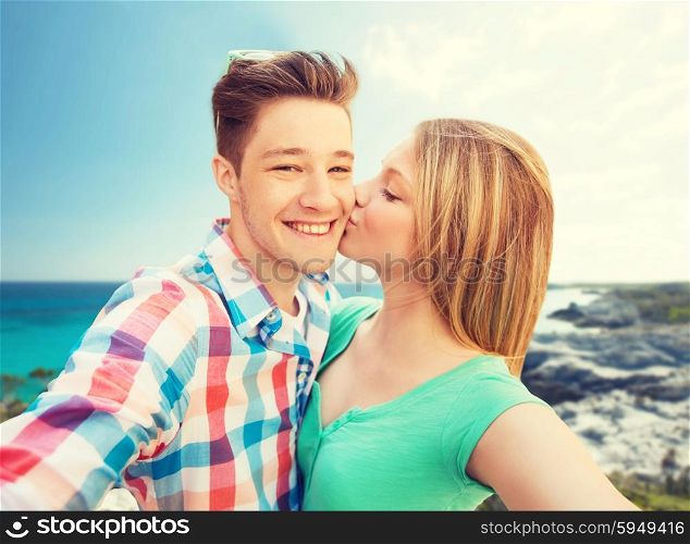 travel, tourism, summer vacation, technology and love concept - happy couple taking selfie with smartphone or camera and kissing over sea shore background