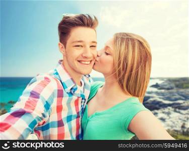 travel, tourism, summer vacation, technology and love concept - happy couple taking selfie with smartphone or camera and kissing over sea shore background