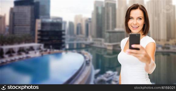 travel, tourism, summer vacation, technology and people - happy young woman taking selfie picture by smartphone over dubai city background